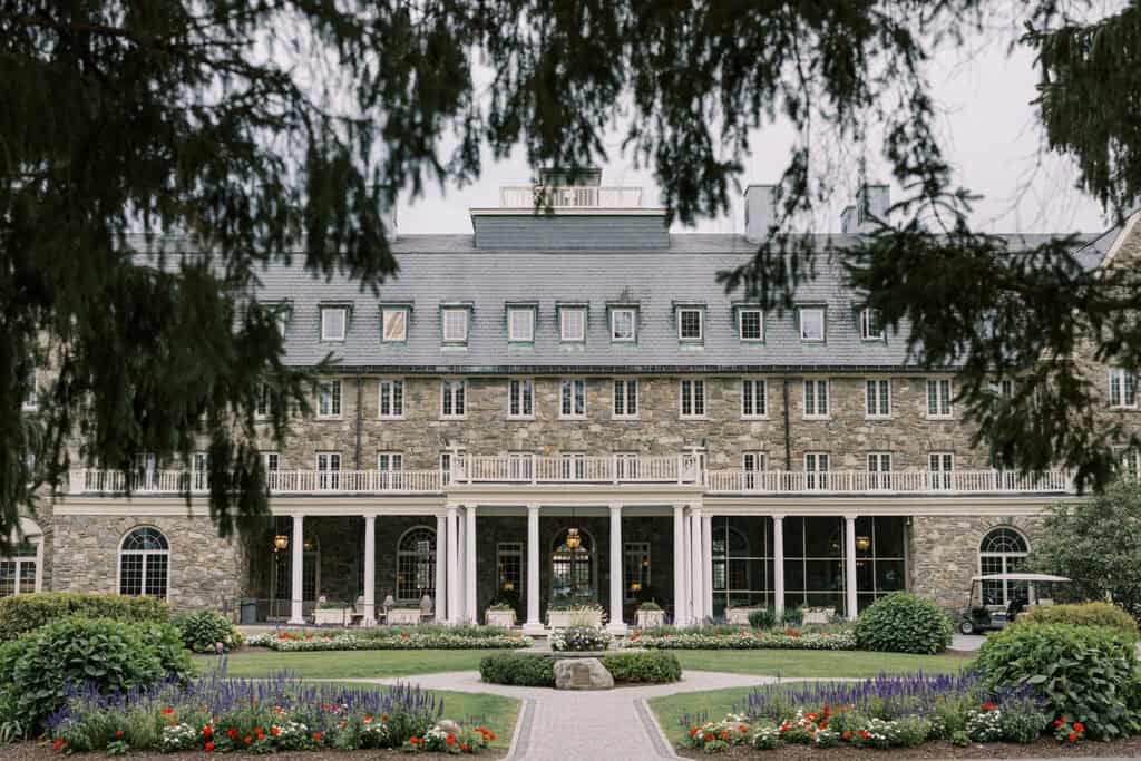 Exterior of Skytop Lodge wedding venue framed by evergreen trees, showing the stone facade, white columned veranda, and manicured gardens in Skytop, Pennsylvania