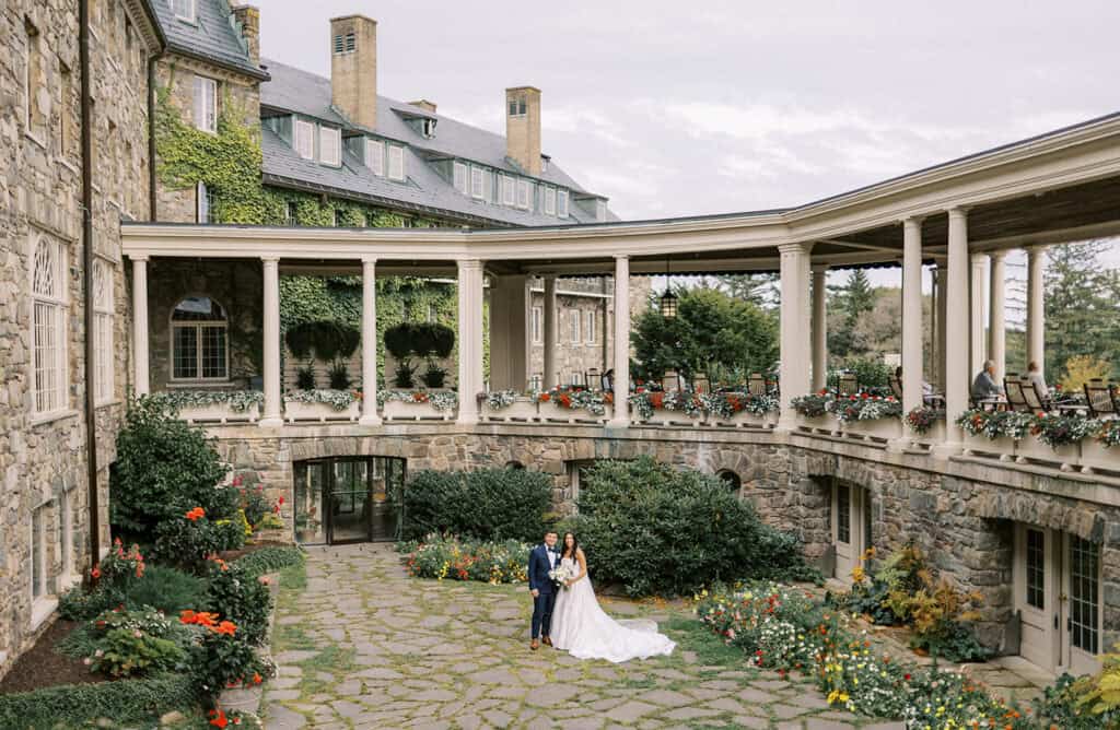 Bride and groom in wedding attire standing in the stone courtyard at Skytop Lodge, surrounded by ivy-covered walls and blooming flower boxes in Skytop, Pennsylvania
