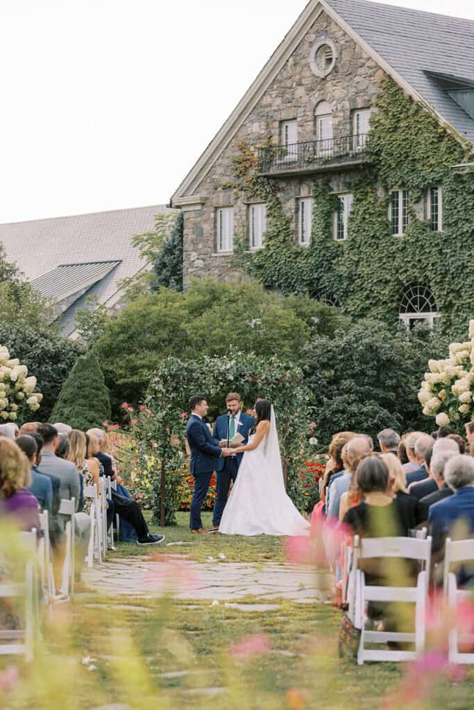 Bride and groom exchanging vows at an outdoor garden ceremony at Skytop Lodge, with guests seated in white chairs and the ivy-covered stone lodge in the background