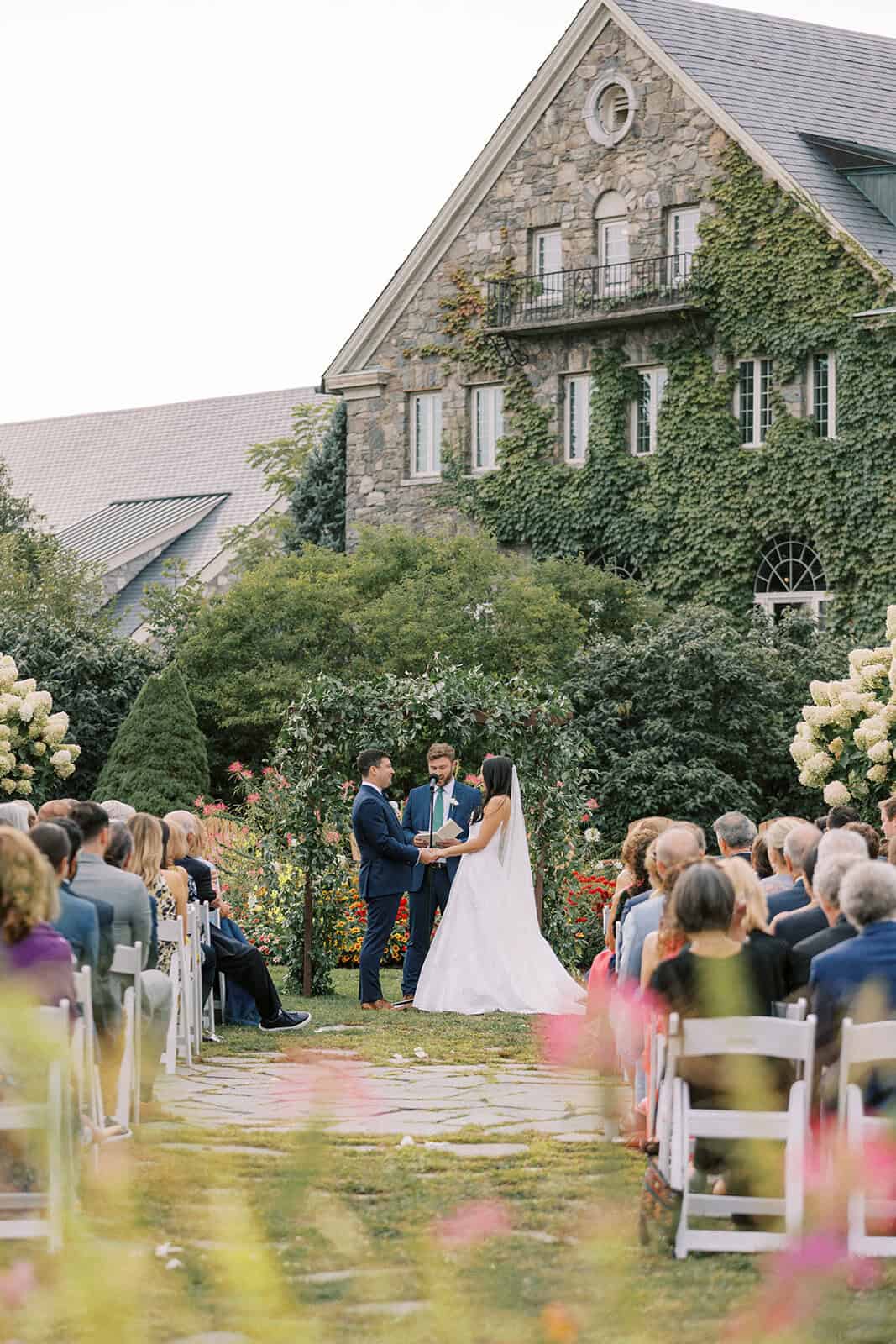 Bride and groom exchanging vows at an outdoor garden ceremony at Skytop Lodge, with guests seated in white chairs and the ivy-covered stone lodge in the background