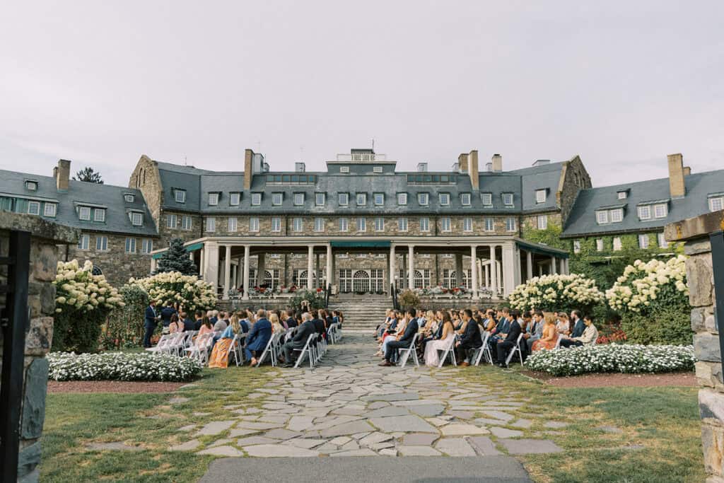 Wide ceremony view of a Skytop Lodge outdoor wedding with guests seated facing the stone lodge, flanked by large blooming hydrangea bushes in the Pocono Mountains