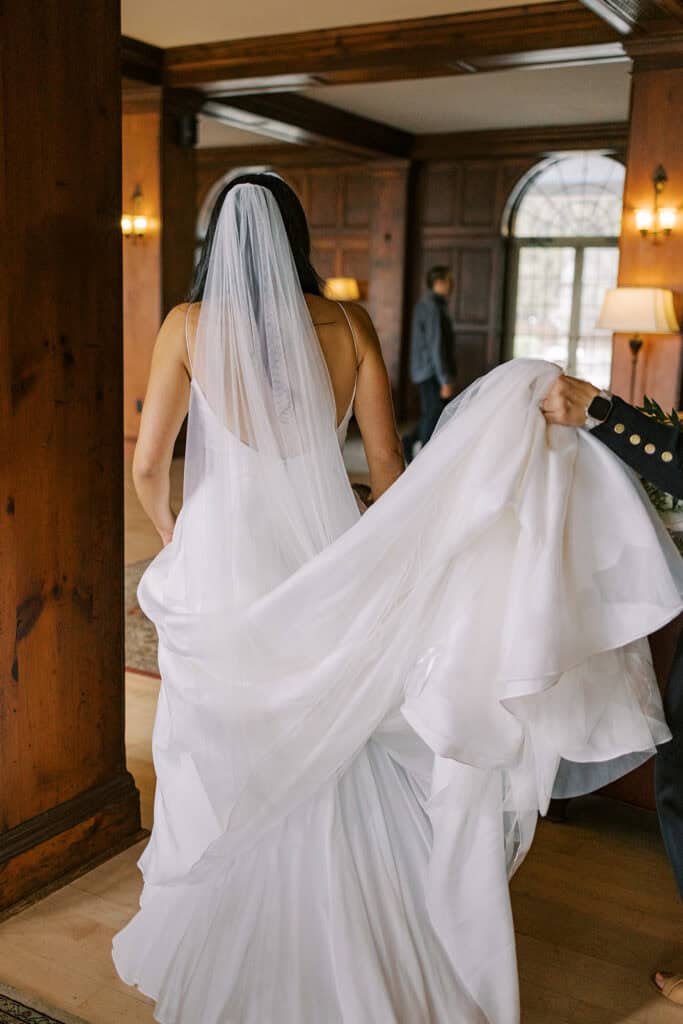 Bride in wedding gown and veil walking through a wood-paneled hallway at Skytop Lodge as her train is carried behind her on wedding day