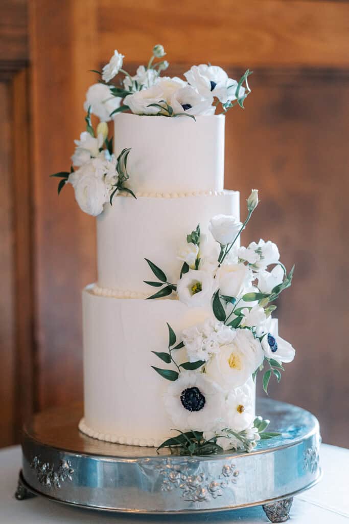 Three-tier white wedding cake decorated with cascading white anemones, ranunculus, and lisianthus on a silver cake stand at a Skytop Lodge wedding reception