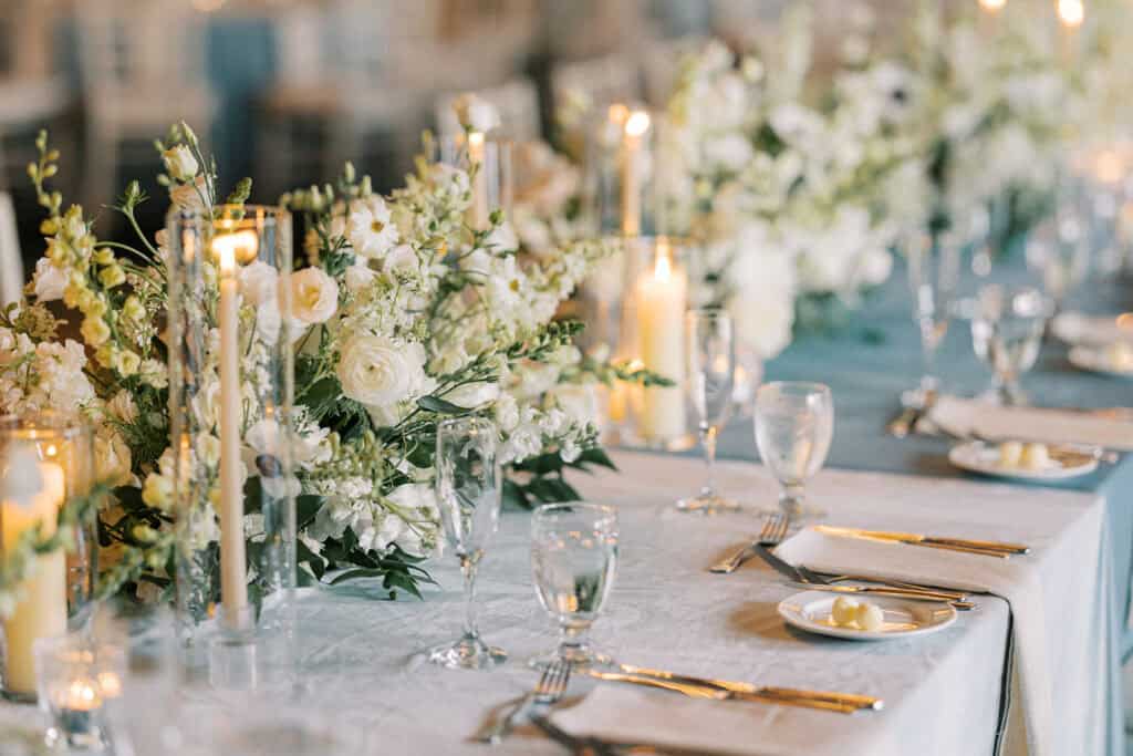 Close-up of white garden floral centerpiece with ranunculus, stock, and greenery alongside taper and pillar candles on a dusty blue linen reception table at Skytop Lodge