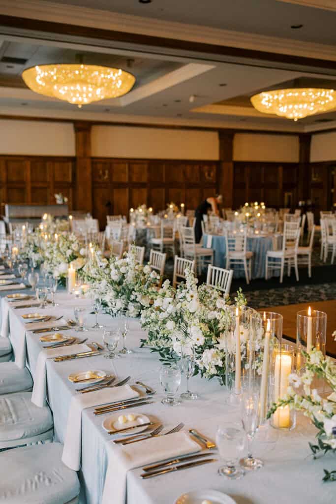Skytop Lodge wedding ballroom reception setup with lush white floral runner centerpieces, taper candles, dusty blue linens, and chandelier lighting in Skytop, Pennsylvania