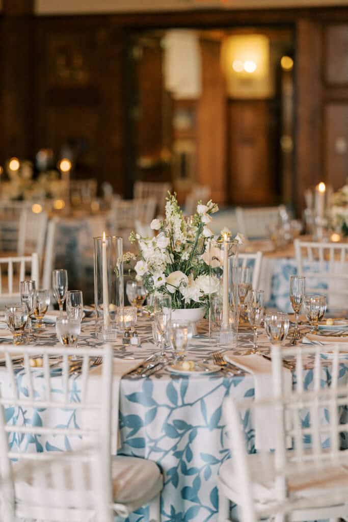 Round reception table detail with blue and white botanical linen, white garden floral centerpiece, taper candles, and chiavari chairs in the wood-paneled ballroom at Skytop Lodge