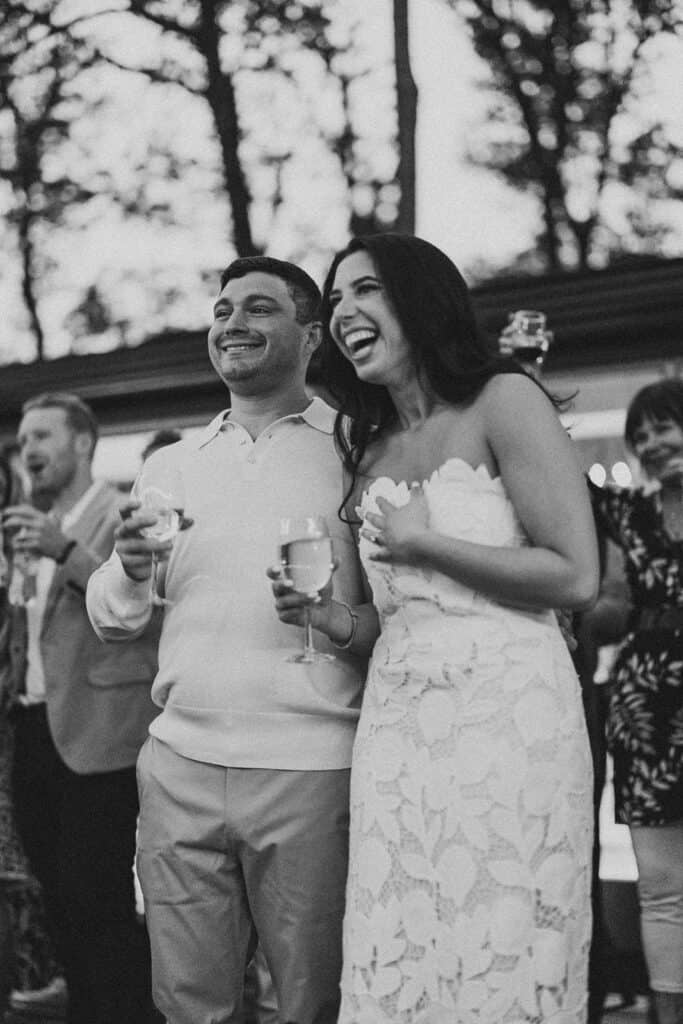 Black and white photo of bride and groom in rehearsal dinner attire laughing together during toasts on the outdoor deck at Skytop Lodge in Skytop, Pennsylvania