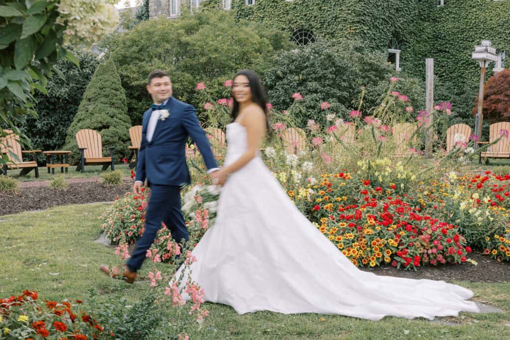 Artistic motion blur wedding day portrait of bride and groom walking hand in hand through the colorful summer gardens in front of the ivy-covered stone lodge at Skytop Lodge in Skytop, Pennsylvania