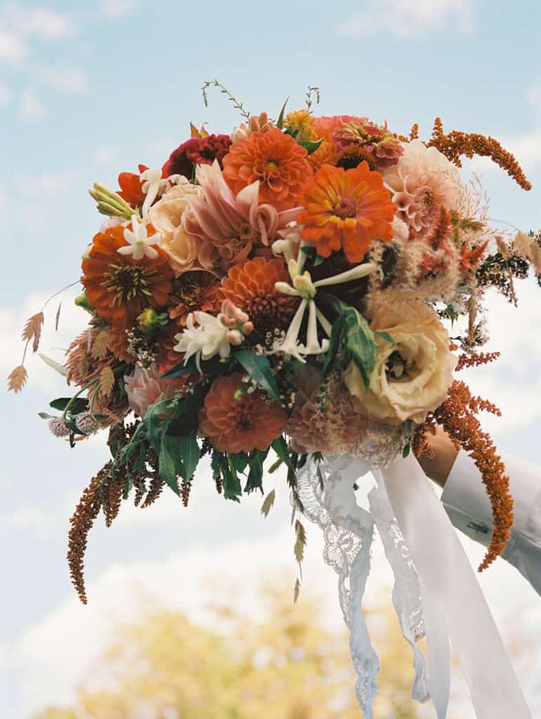 Fall bridal bouquet of orange dahlias, zinnias, burgundy blooms, cream lisianthus, and lace ribbon held up against a blue sky at Tyler Arboretum September wedding, photographed on film