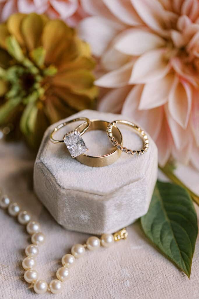 Close-up detail of cushion cut diamond engagement ring, gold wedding bands, and pearl bracelet in a velvet ring box surrounded by blush and peach dahlias, Tyler Arboretum fall wedding