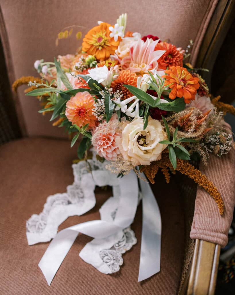 Fall bridal bouquet by Rustic Bunch resting on a velvet chair — orange and blush dahlias, zinnias, cream lisianthus, and trailing lace ribbon, Tyler Arboretum September wedding