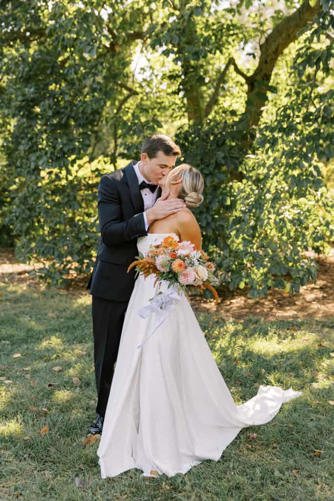 Bride and groom sharing a tender kiss in the gardens at Tyler Arboretum, bride in open back strapless gown with cathedral train and fall bouquet, Media Pennsylvania fall wedding photography