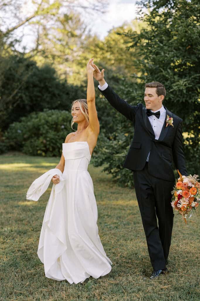 Bride and groom laughing while groom spins bride in the Tyler Arboretum gardens, groom holding fall bouquet of orange dahlias and zinnias, Philadelphia fall wedding photography