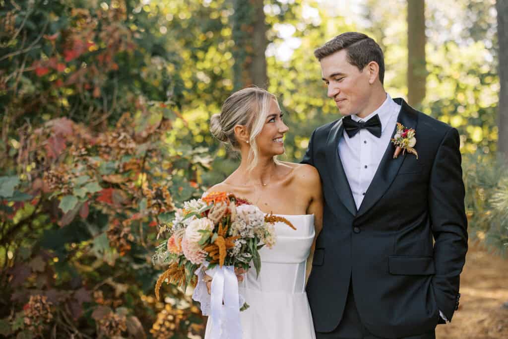 Bride smiling up at groom surrounded by vibrant fall foliage at Tyler Arboretum, bride in strapless gown holding lush fall bouquet by Rustic Bunch, Media Pennsylvania wedding photographer Samantha Jay Photo
