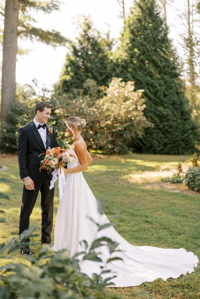 Bride and groom portrait framed by garden greenery and evergreen trees at Tyler Arboretum, bride's cathedral train trailing across the lawn, fall wedding photography near Philadelphia