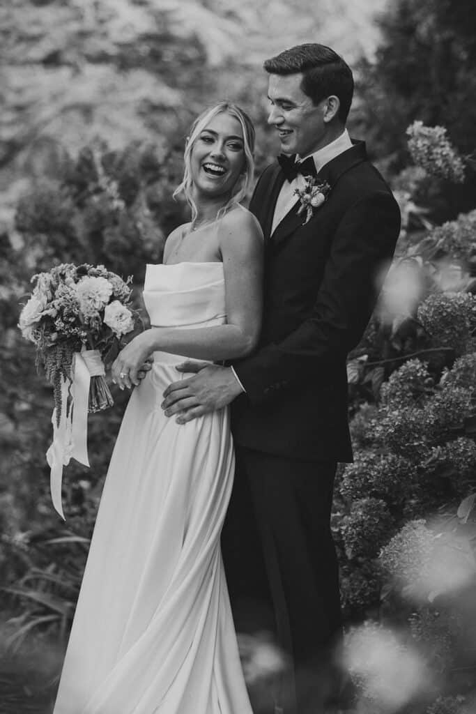 Black and white couple portrait of bride laughing joyfully with groom embracing her beside hydrangeas in the Tyler Arboretum gardens, Philadelphia fall wedding photographer Samantha Jay Photo