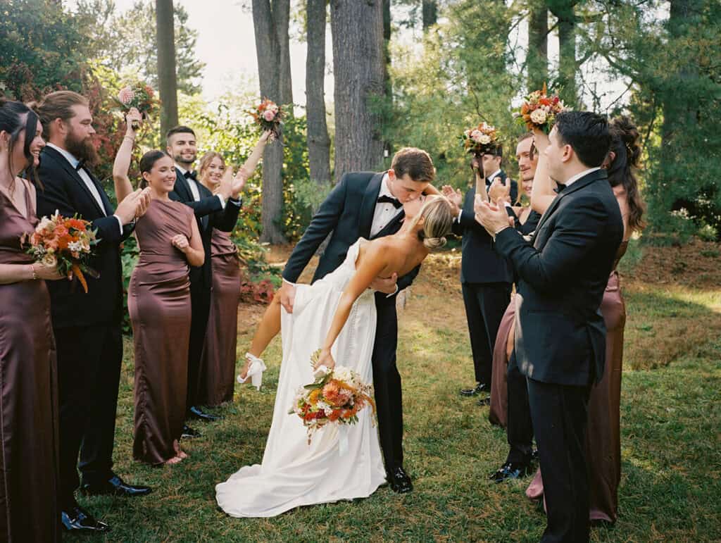 Groom dipping bride for a kiss while full wedding party cheers around them in the pine grove at Tyler Arboretum, bridesmaids in chocolate brown satin gowns, fall wedding photography Media PA
