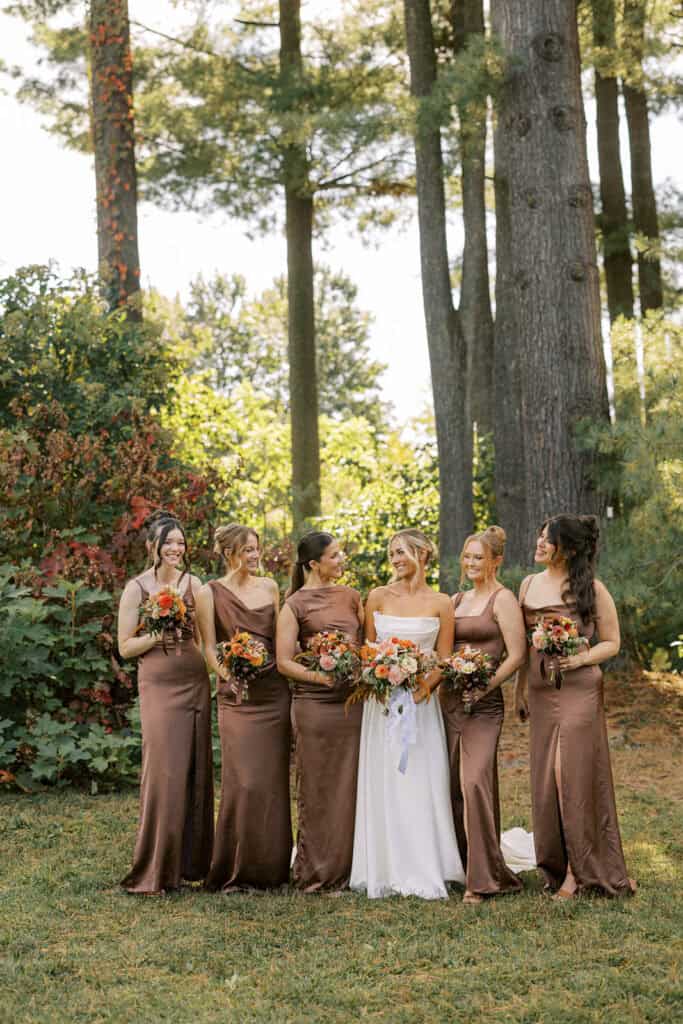 Bride with five bridesmaids in chocolate brown satin gowns holding fall bouquets of orange dahlias and zinnias in the gardens at Tyler Arboretum, September wedding photography near Philadelphia