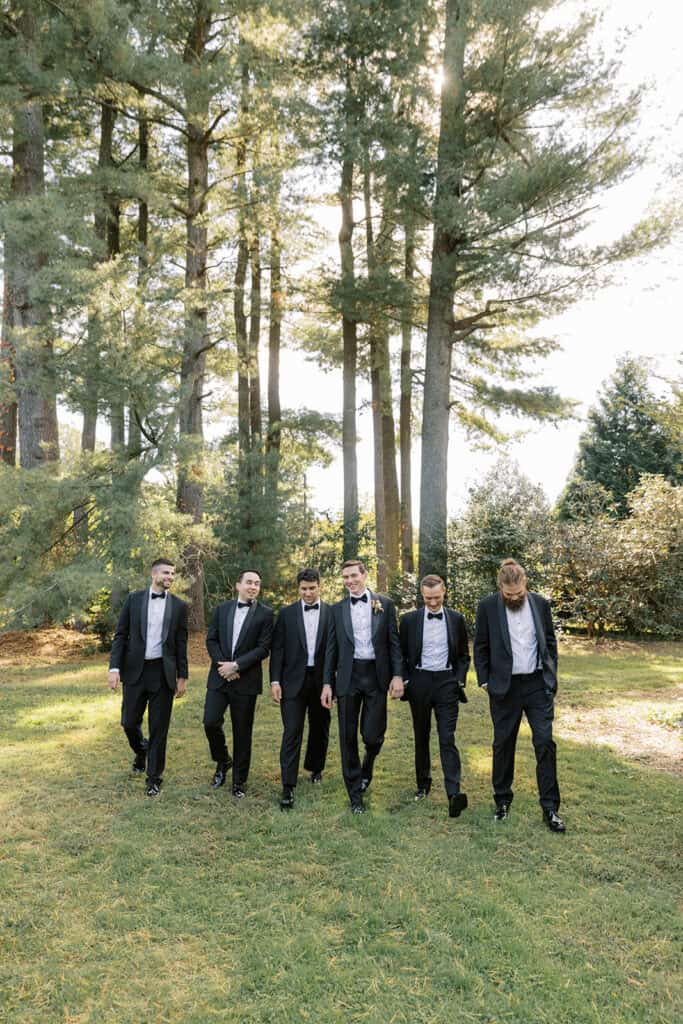 Groom and five groomsmen in black tuxedos walking together through the pine grove at Tyler Arboretum fall wedding, Media Pennsylvania, photographed by Samantha Jay Photo