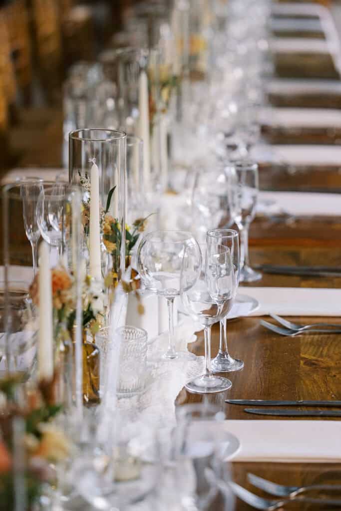 Long farm table at Tyler Arboretum wedding reception with white lace runner, tall taper candles in hurricane vases, and fall floral centerpieces, Media PA