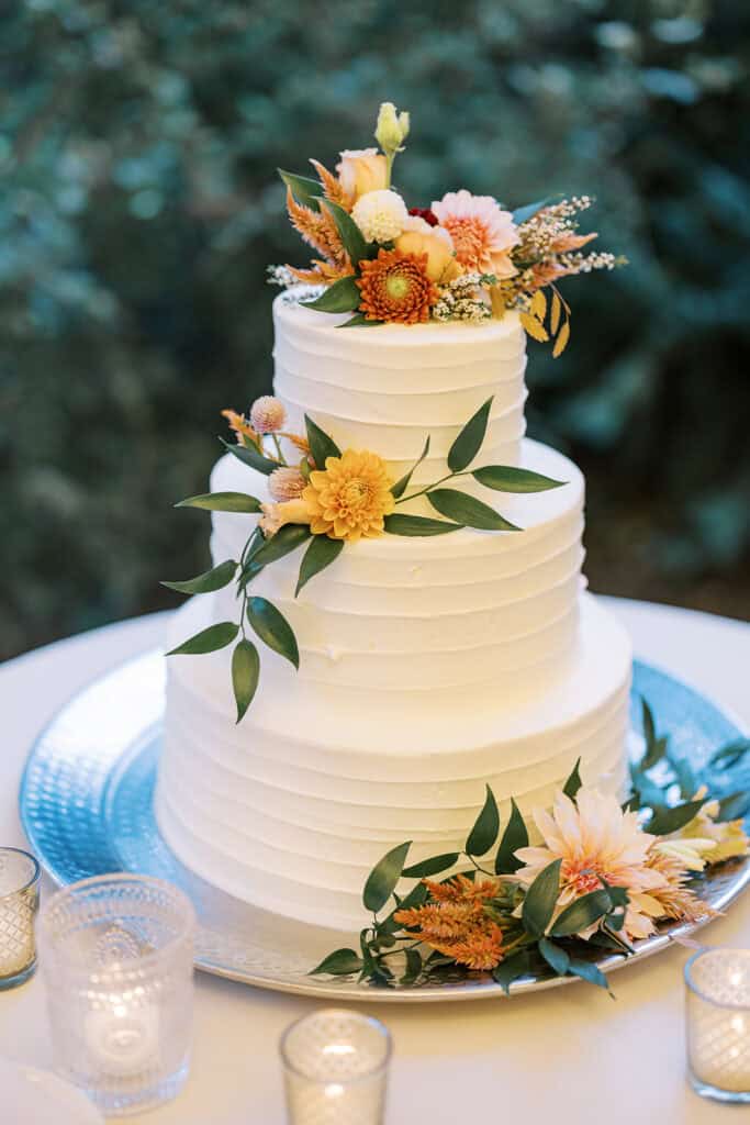 Three-tier white buttercream wedding cake decorated with orange dahlias, mums, blush roses, and greenery at Tyler Arboretum fall wedding reception