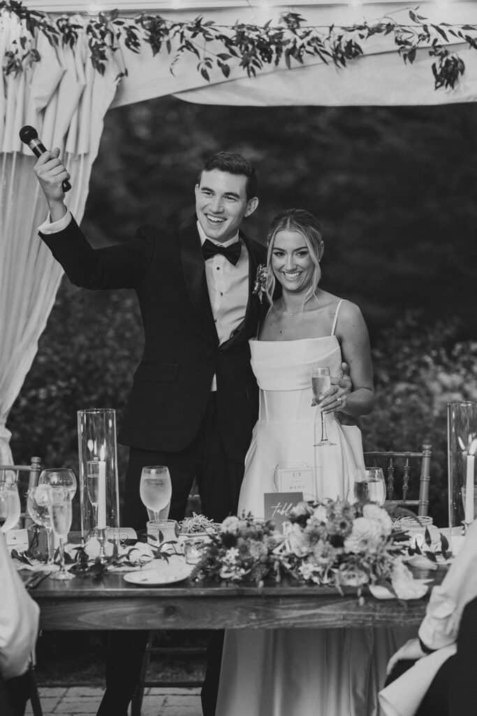 Black and white photo of groom raising a microphone during welcome toast with bride at their sweetheart table, Tyler Arboretum wedding reception