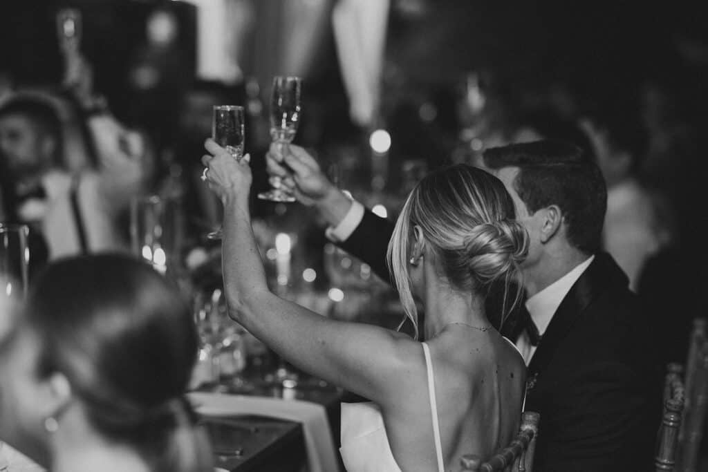 Black and white detail of bride raising her champagne flute during toasts at Tyler Arboretum wedding reception, bride's low chignon updo visible from behind