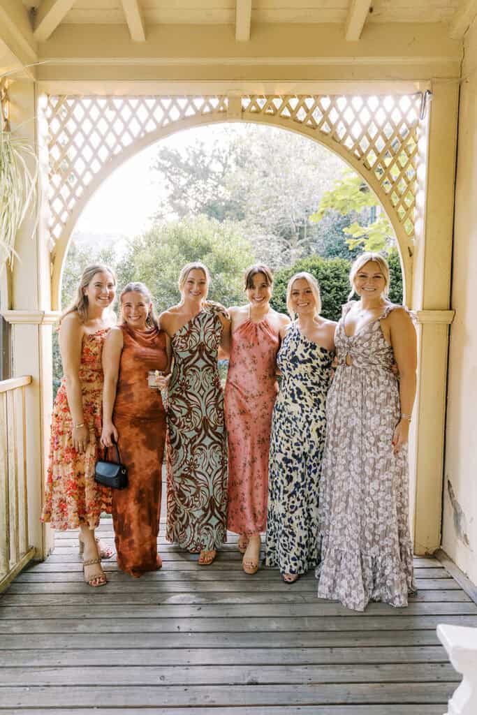 Group of wedding guests in colorful floral and printed maxi dresses posed under a white lattice arch at Tyler Arboretum during fall wedding cocktail hour