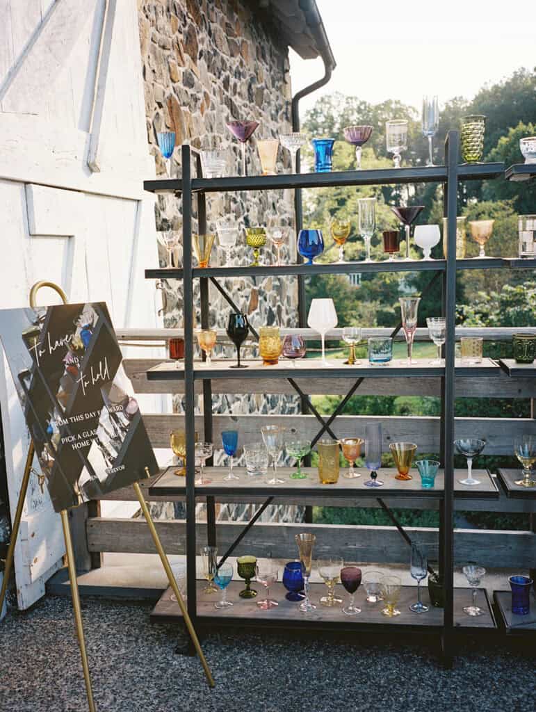 Colorful vintage glassware wedding favor display on a rustic metal shelving unit outside Tyler Arboretum, with a handwritten "pick a glass to take home" sign