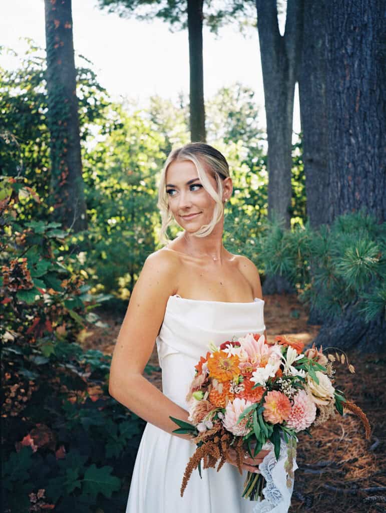 Film portrait of bride in strapless white gown holding an orange and peach fall bouquet with lace ribbon among autumn trees at Tyler Arboretum