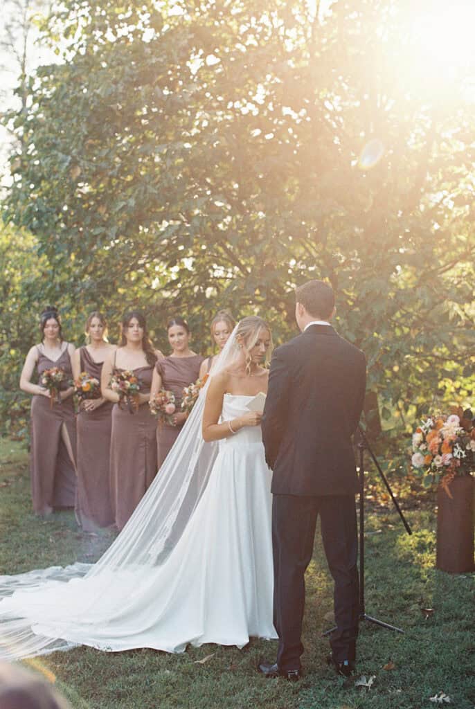 Outdoor wedding ceremony in Tyler Arboretum's Magnolia Garden, bride reading vows to groom with bridesmaids in chocolate brown gowns, golden September light