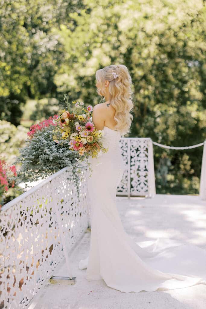 Bride in Madi Lane off-shoulder gown holding wildflower bouquet on wrought iron balcony overlooking gardens at Drumore Estate wedding venue Lancaster PA