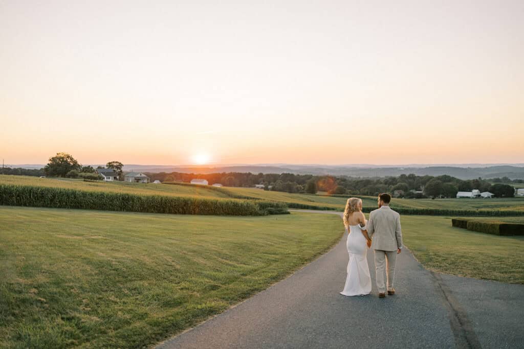 Bride and groom walking hand in hand toward Lancaster County sunset on country road at Drumore Estate summer wedding in Pequea Pennsylvania