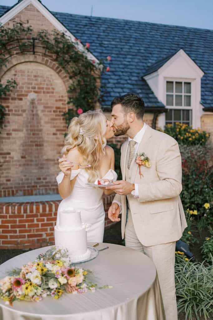Bride and groom kissing after cake cutting at Drumore Estate summer wedding with wildflower arrangements and brick carriage house backdrop