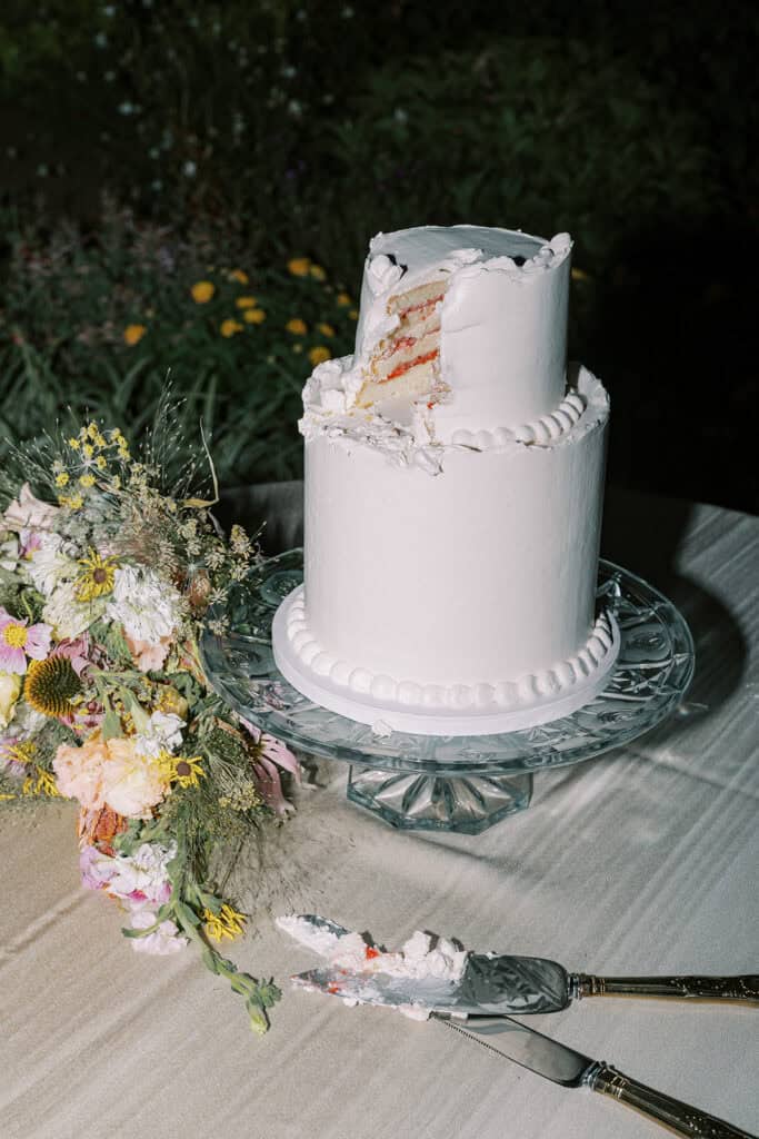 Sliced two-tier white wedding cake by Salt and Light Bakery on glass cake stand with wildflower bouquet at Drumore Estate Lancaster PA