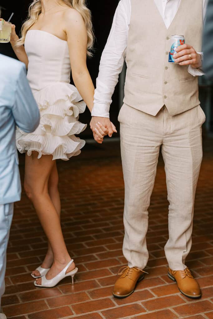 Bride in ruffle mini reception dress holding hands with groom in tan linen vest on dance floor at Drumore Estate summer wedding
