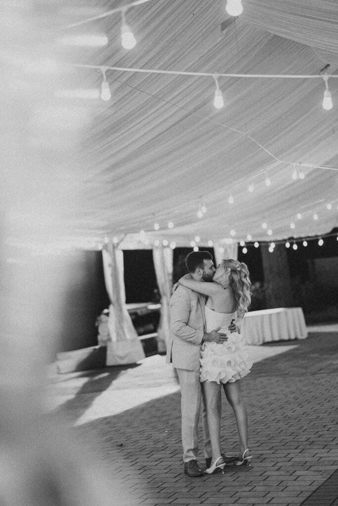 Black and white film photo of bride and groom kissing under string lights inside Drumore Estate reception tent at summer wedding