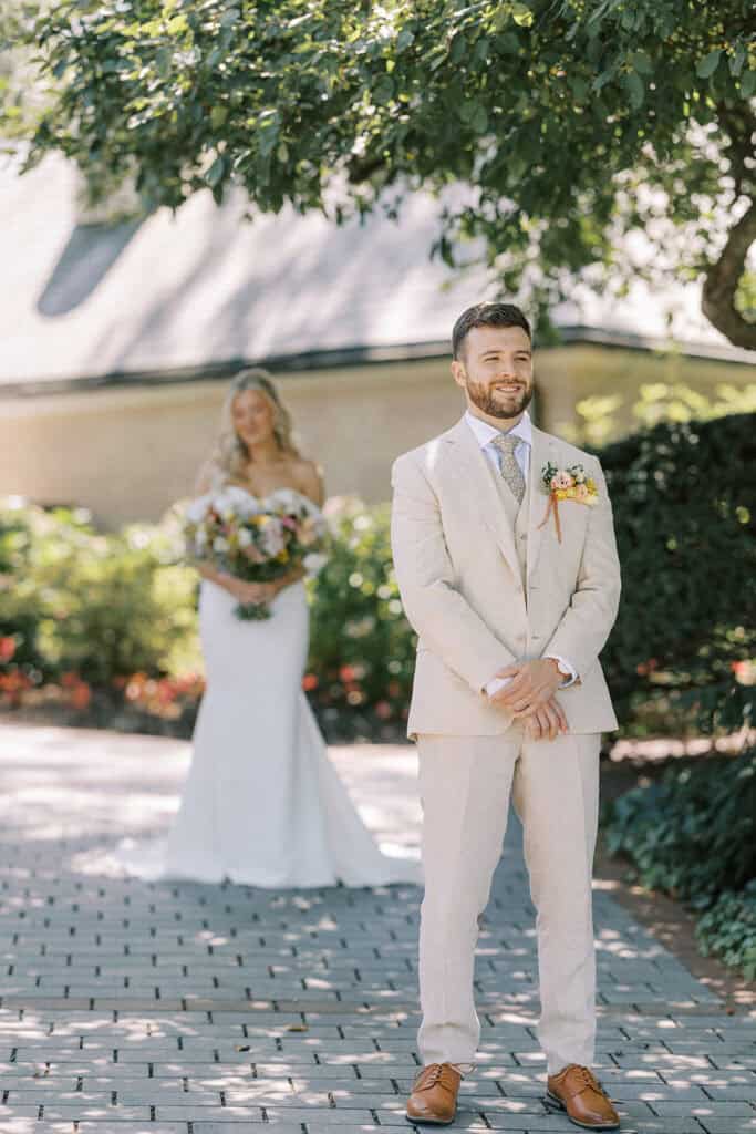 Groom in a tan linen suit smiling during first look at Drumore Estate wedding in Pequea PA with bride in Madi Lane gown approaching behind him