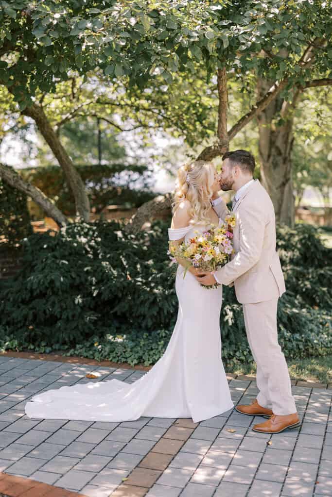Bride and groom kissing under summer trees at Drumore Estate Lancaster PA wedding holding colorful wildflower bouquet