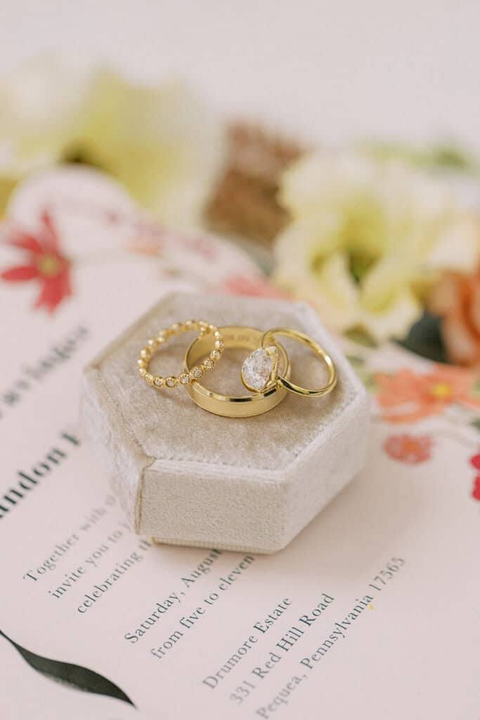 Gold oval engagement ring and wedding bands in velvet hexagon ring box atop floral Drumore Estate wedding invitation Lancaster Pennsylvania