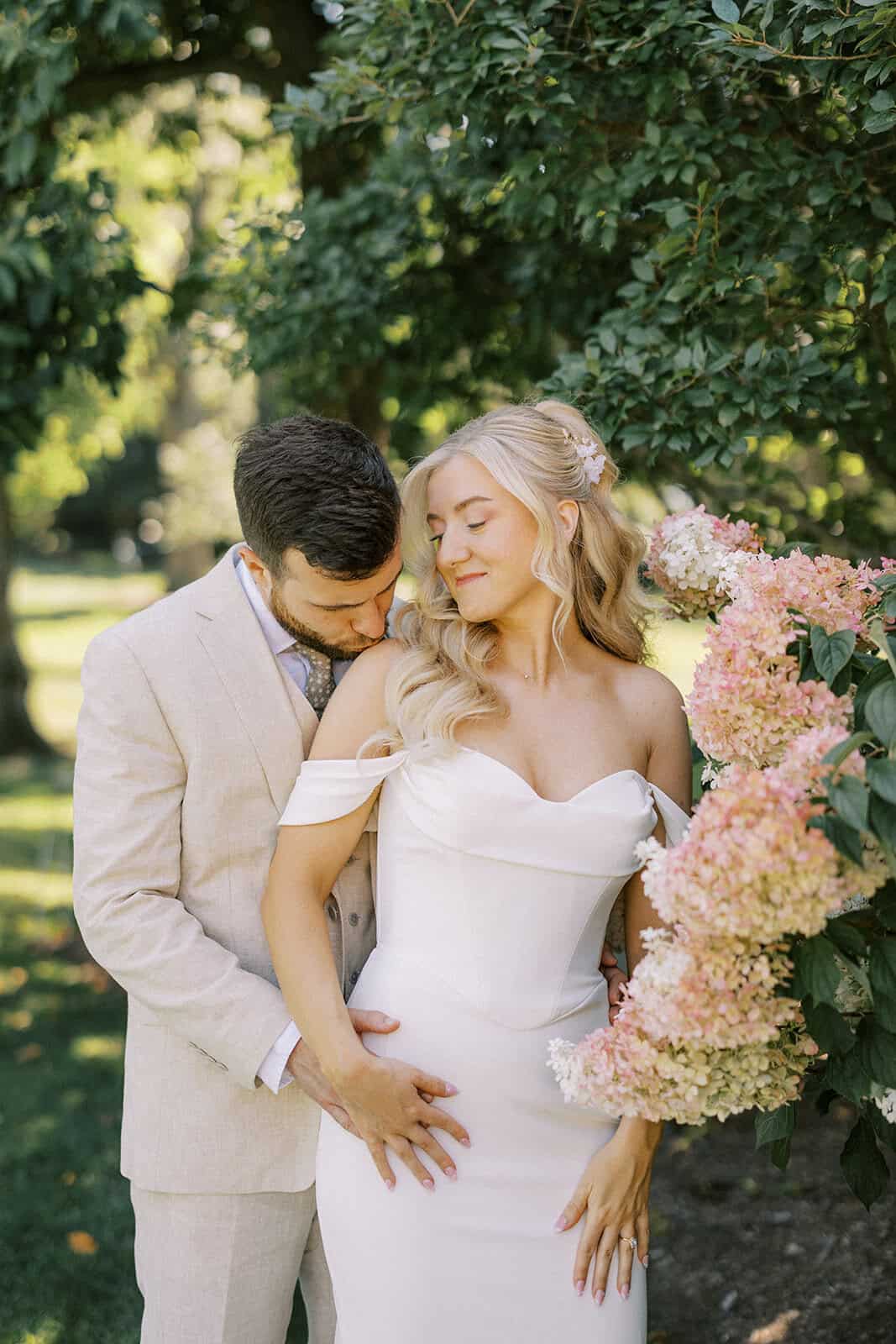 Groom kissing bride's shoulder beside blooming pink hydrangeas at outdoor Drumore Estate summer wedding