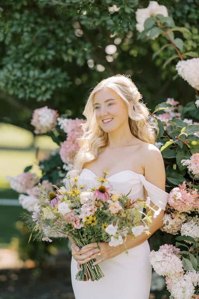 Bride in off-shoulder Madi Lane gown smiling with wildflower bouquet surrounded by pink hydrangeas at Drumore Estate wedding venue