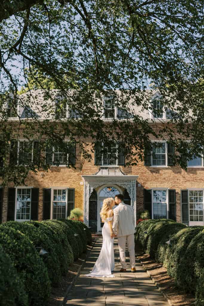 Bride and groom from behind walking toward Drumore Estate mansion entrance lined with trimmed boxwood hedges at summer wedding