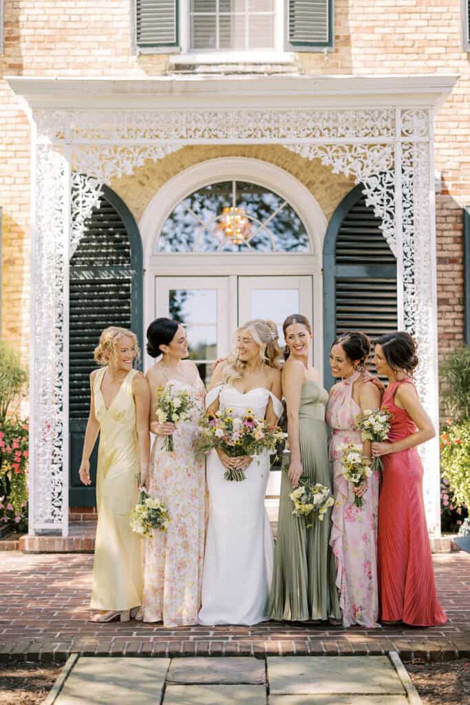 Bride and five bridesmaids in mixed color summer dresses with wildflower bouquets standing in front of ornate white ironwork entrance at Drumore Estate wedding venue