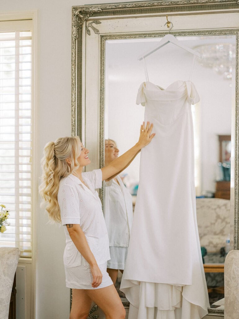 Bride smiling and reaching for her Madi Lane off-shoulder gown hanging on ornate mirror in Drumore Estate bridal suite Lancaster PA