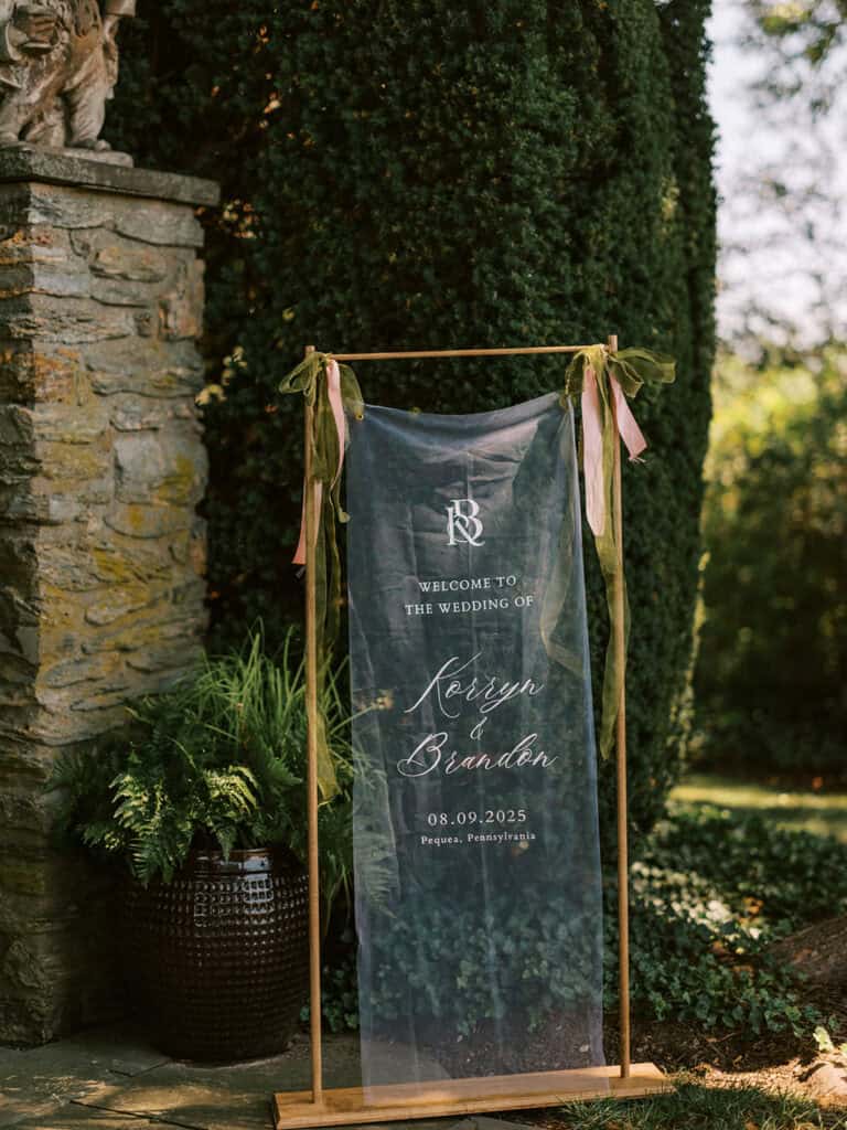 Sheer fabric wedding welcome sign for Korryn and Brandon on wooden frame with green and pink ribbon ties in Drumore Estate garden Pequea PA