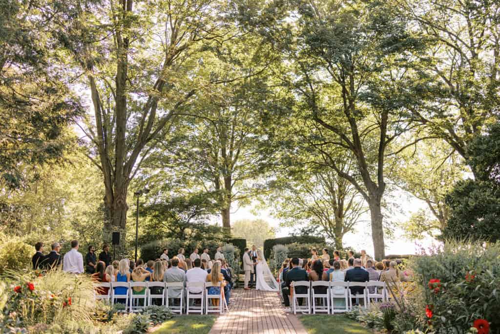 Wide shot of outdoor summer wedding ceremony at Drumore Estate Lancaster PA with guests seated among formal gardens and tall shade trees