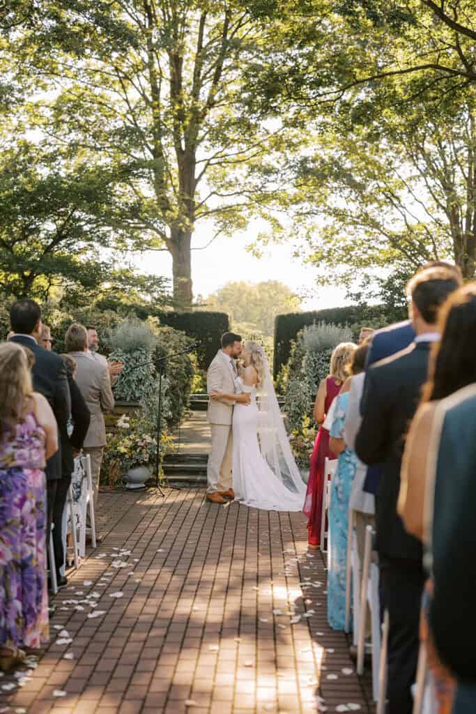 Bride and groom first kiss at outdoor ceremony at Drumore Estate wedding venue in Pequea Pennsylvania with rose petal lined brick aisle