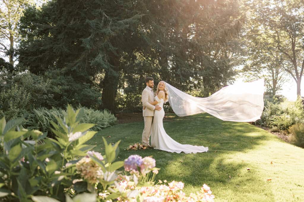 Bride in Madi Lane gown with cathedral veil blowing in wind standing with groom in Drumore Estate formal gardens at summer wedding in Pennsylvania