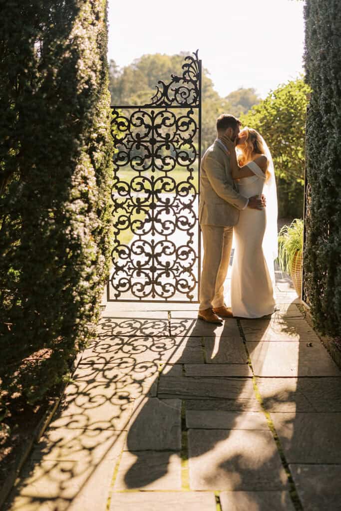 Bride and groom kissing in front of ornate iron garden gate at Drumore Estate wedding venue in Pequea PA during late summer golden hour